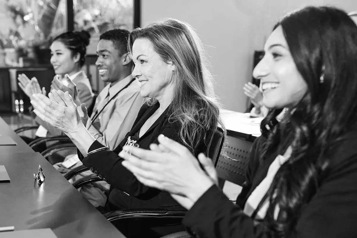 A group of four people are seated in chairs around a table, attentively clapping and looking towards the front of the room. They appear to be in an Australian workplace, actively engaging in a DEI-focused presentation or meeting.