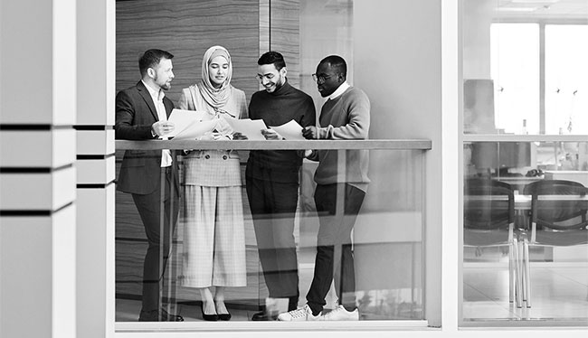 Four professionals stand in a group in the lobby of a corporate office building. The team is reviewing paperwork related to leadership development and executive coaching for succession planning.