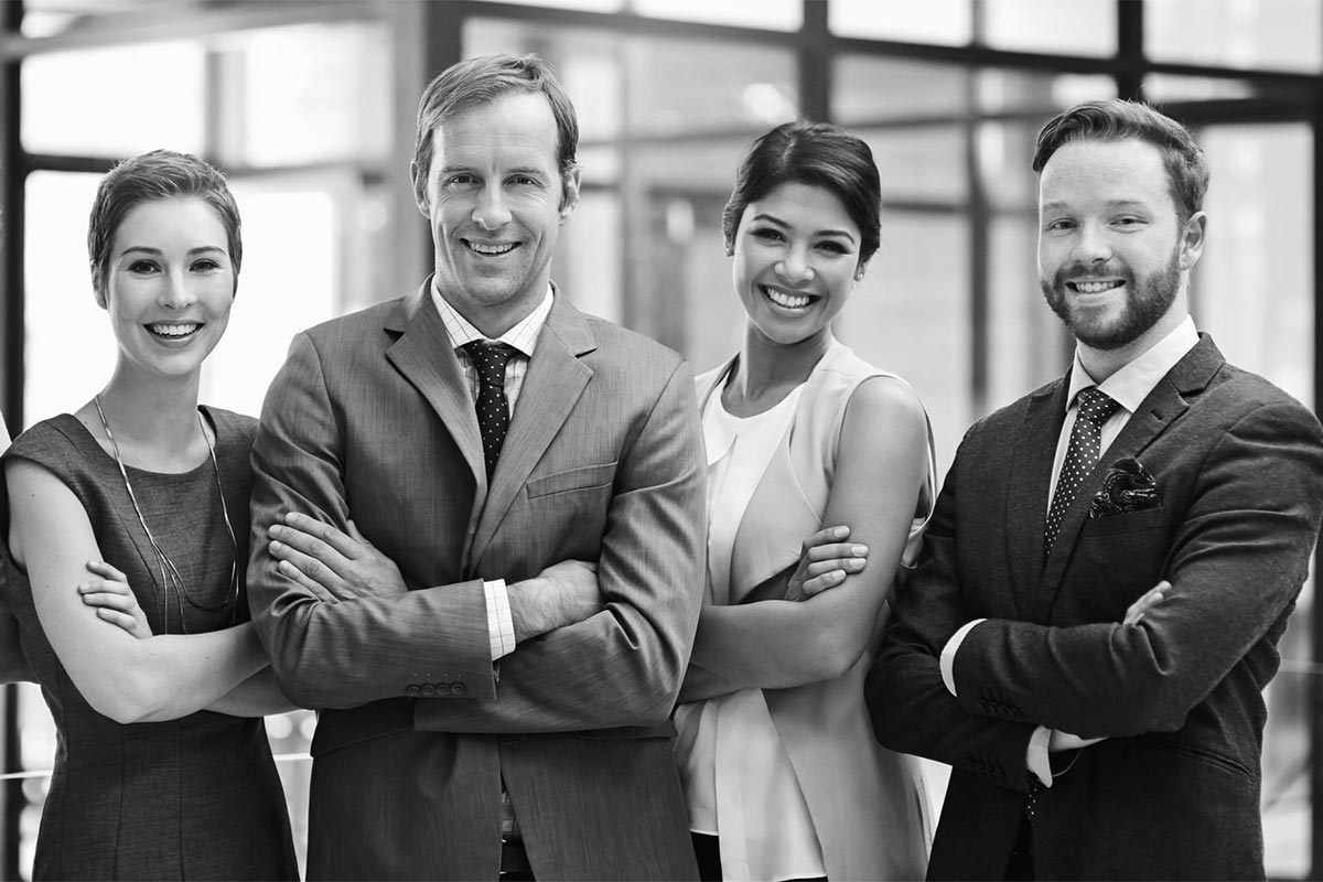 A black and white photo shows four smiling individuals standing side by side with arms crossed, dressed in business attire, against a blurred office background. The group consists of two men and two women, exuding confidence and professionalism, embodying the values of an Australian workplace committed to diversity, equity and inclusion.